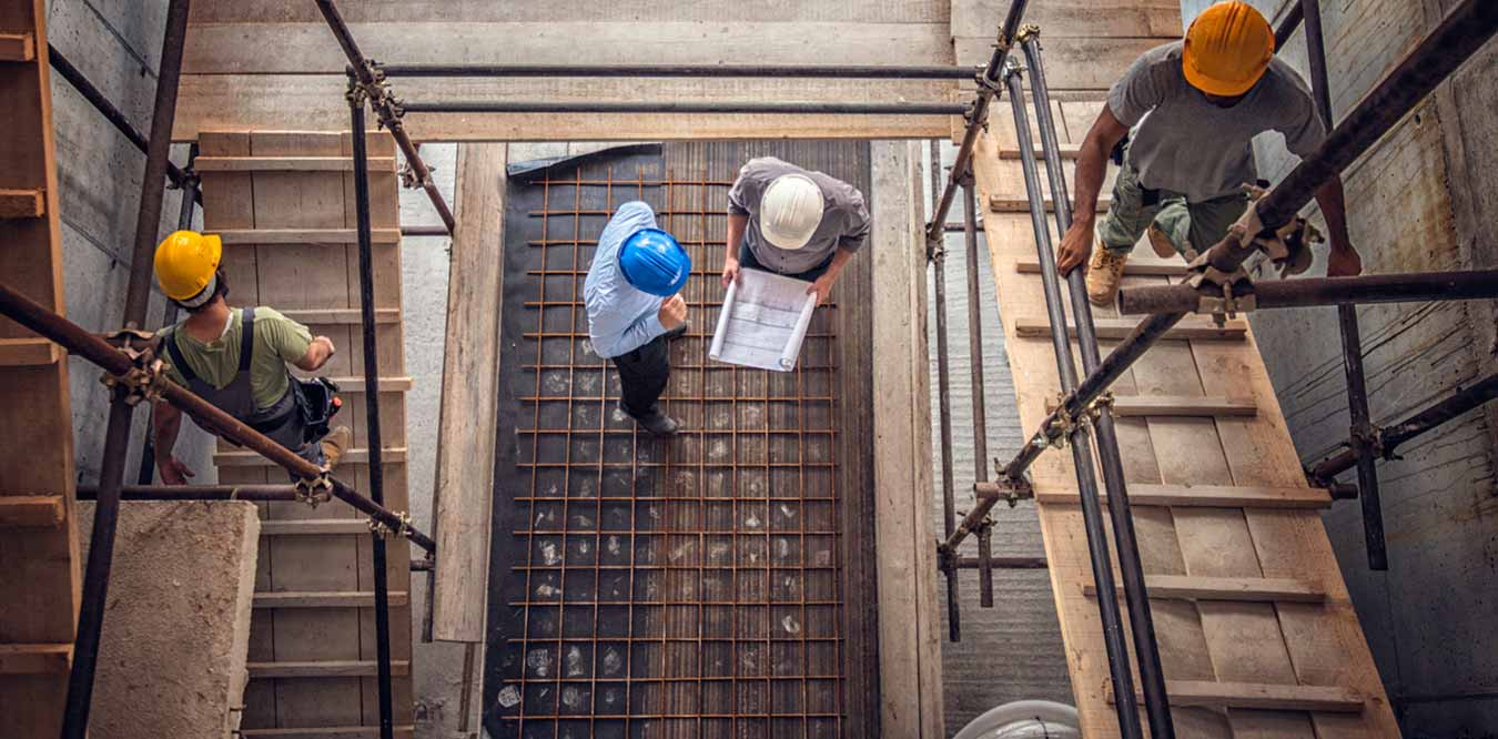 two project managers discussing blueprints near construction workers under scaffolding